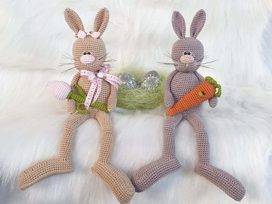 Zwei gehäkelte Amigurumi-Hasen mit Karotte und Blume vor Osternest auf weißem Fell