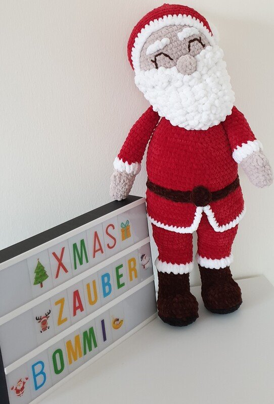 Gehäkelter Amigurumi‑Weihnachtsmann mit rotem Anzug und weißem Bart neben einer Leuchtbox mit bunten Buchstaben