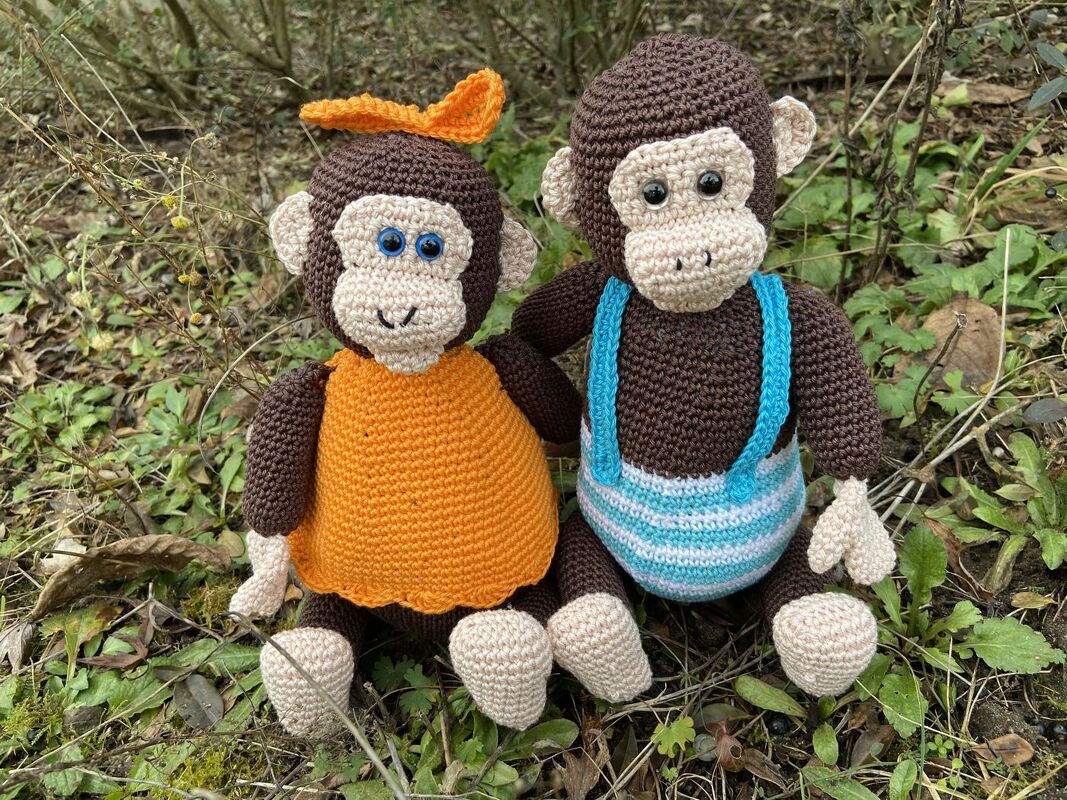 Zwei gehäkelte Amigurumi-Affen im Wald, einer mit orangefarbenem Kleid und Schleife, der andere mit blau-weiß gestreifter Latzhose
