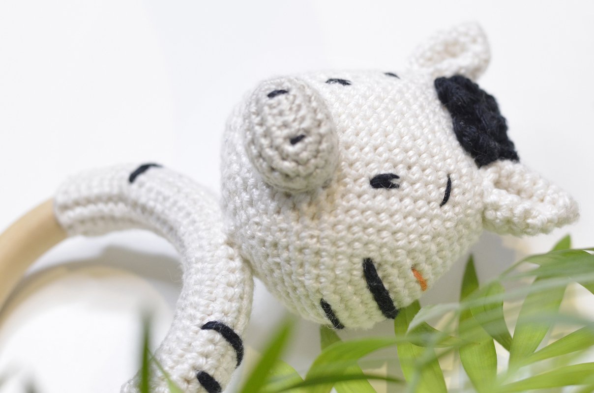 Gehäkelter weißer Greifring mit Tierkopf, schwarzen Details und hellem Holzring neben grünen Blättern.