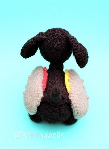 Back view of a dark brown crocheted hot dog amigurumi with floppy ears, short tail, and beige bun-like side panels with pink and yellow condiment accents on a light aqua background.