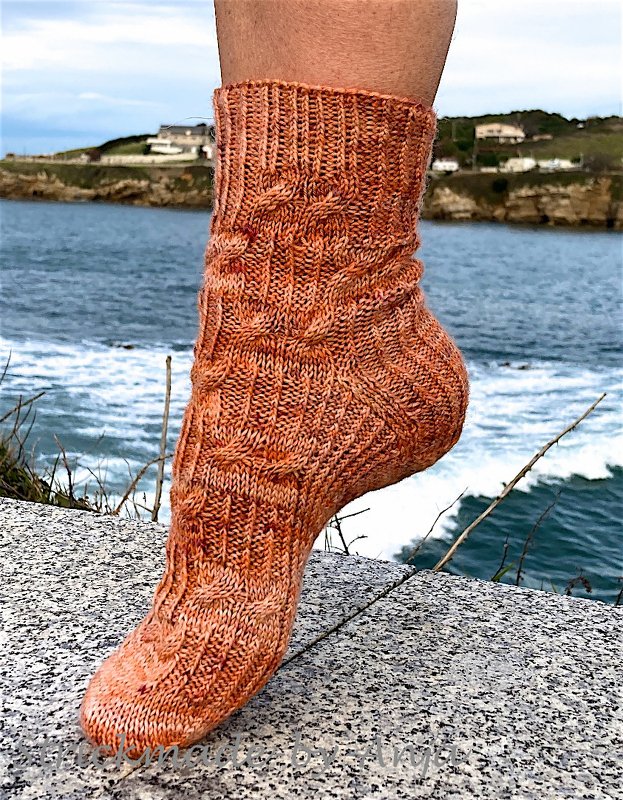 Seitliche Nahaufnahme einer orange-braunen handgestrickten Damensocke mit Zopf- und Rippenmustern, vor Küstenhintergrund