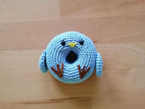Hellblau gehäkeltes Donut‑Vögelchen mit Flügeln, schwarzen Augen und gelbem Schnabel auf Holzuntergrund