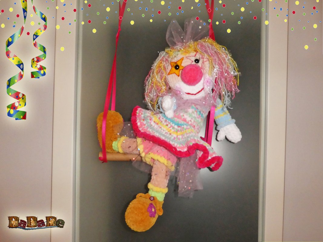 Gehäkelte Clown-Puppe mit buntem, glitzerndem Haar und roter Nase auf einer rosa Schaukel, mit Konfetti und Luftschlangen