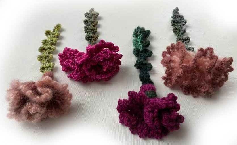 Vier kleine gehäkelte Blumen in Rosa, Magenta und Grün, flach auf weißem Untergrund ausgelegt.