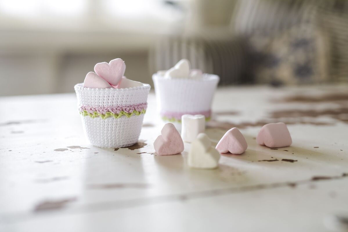 Zwei weiße gehäkelte Mini-Becher mit rosa-grüner Bordüre und rosa sowie weißen herzförmigen Bonbons auf einem hellen Tisch