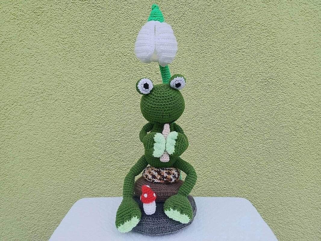 Gehäkelter grüner Amigurumi-Frosch mit großen Augen, weißem Schneeglöckchen auf dem Kopf, hellgrünem Schmetterling in den Händen und kleinem roten Pilz auf grauem Sockel.