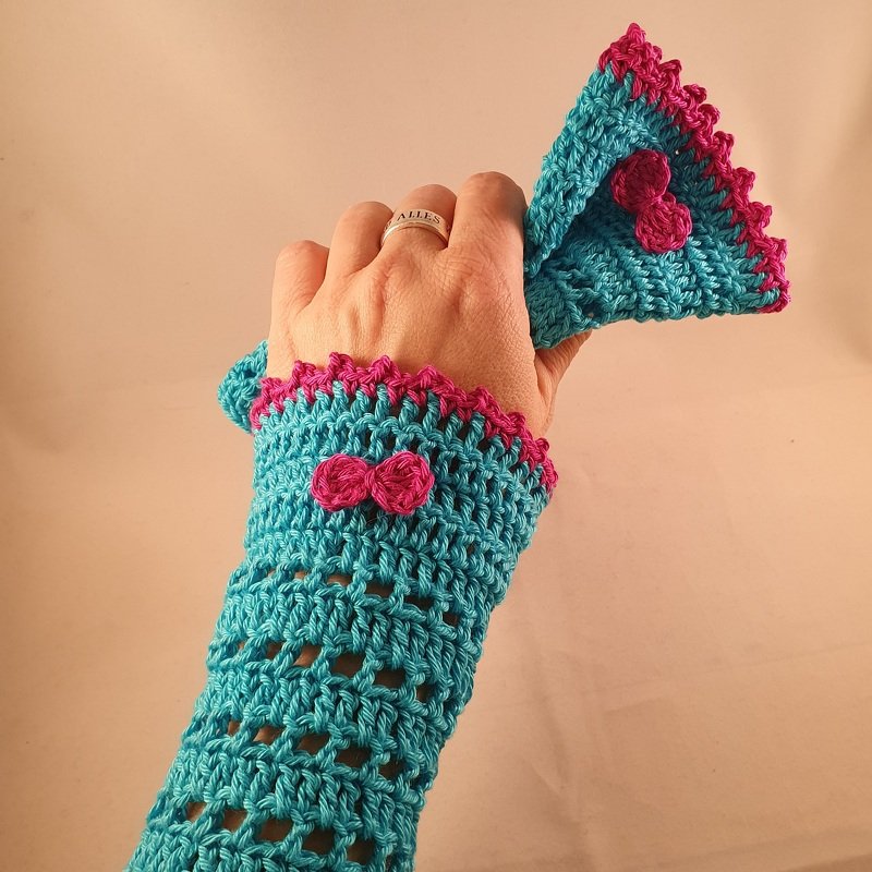 Hand mit türkis-pinken gehäkelten Armstulpen und Schleifen-Applikation