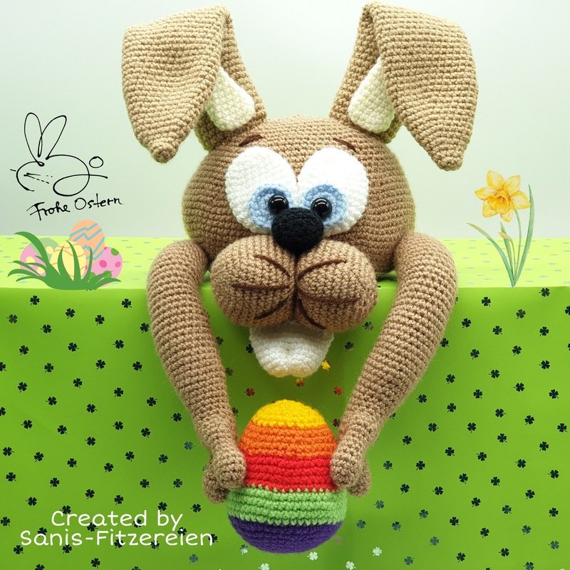 Brauner gehäkelter Osterhase mit schlaffen Ohren und großen Augen hält ein bunt gestreiftes Regenbogen-Osterei.