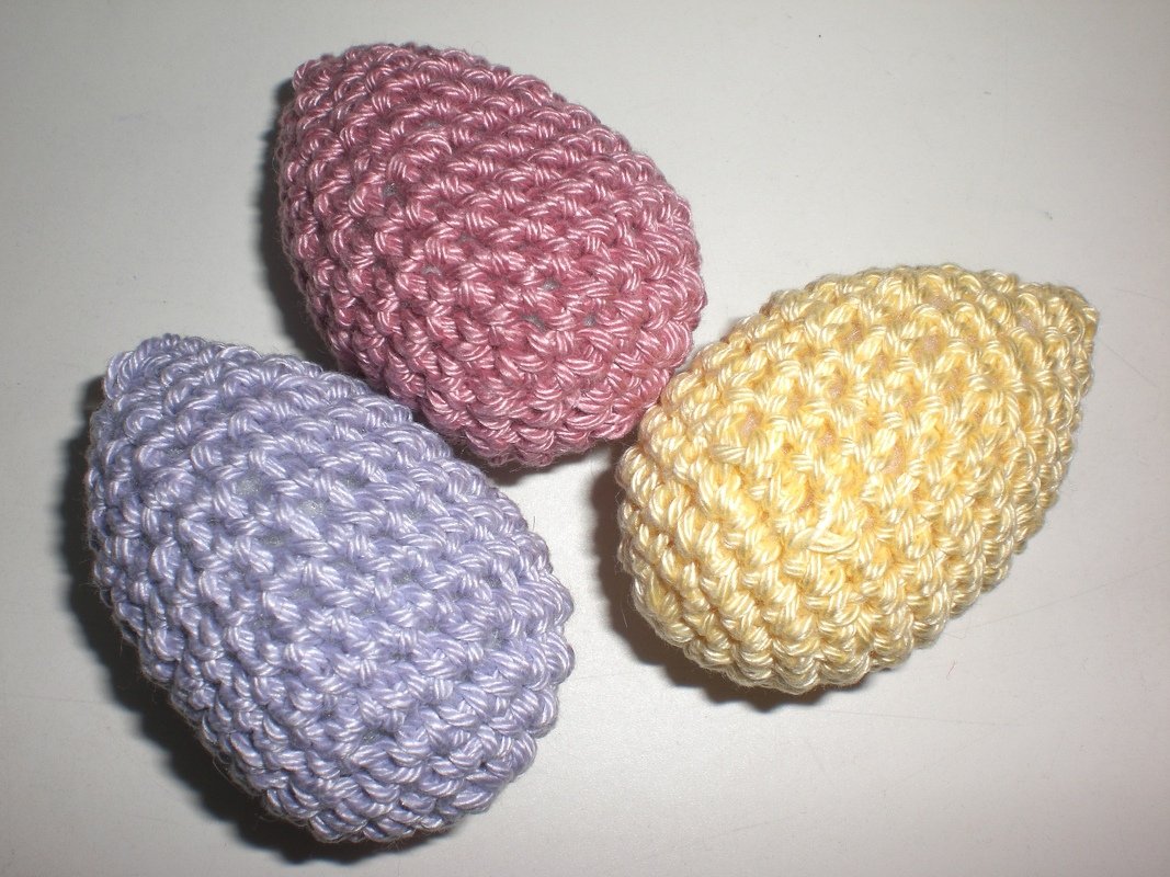 Drei handgehäkelte Ostereier in Pastellfarben (lila, rosa, gelb) auf hellem Untergrund