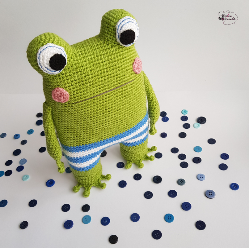Grüne gehäkelte Amigurumi-Froschfigur mit großen Augen, rosigen Wangen und blau-weiß gestreifter Hose auf hellem Untergrund mit verstreuten blauen Knöpfen
