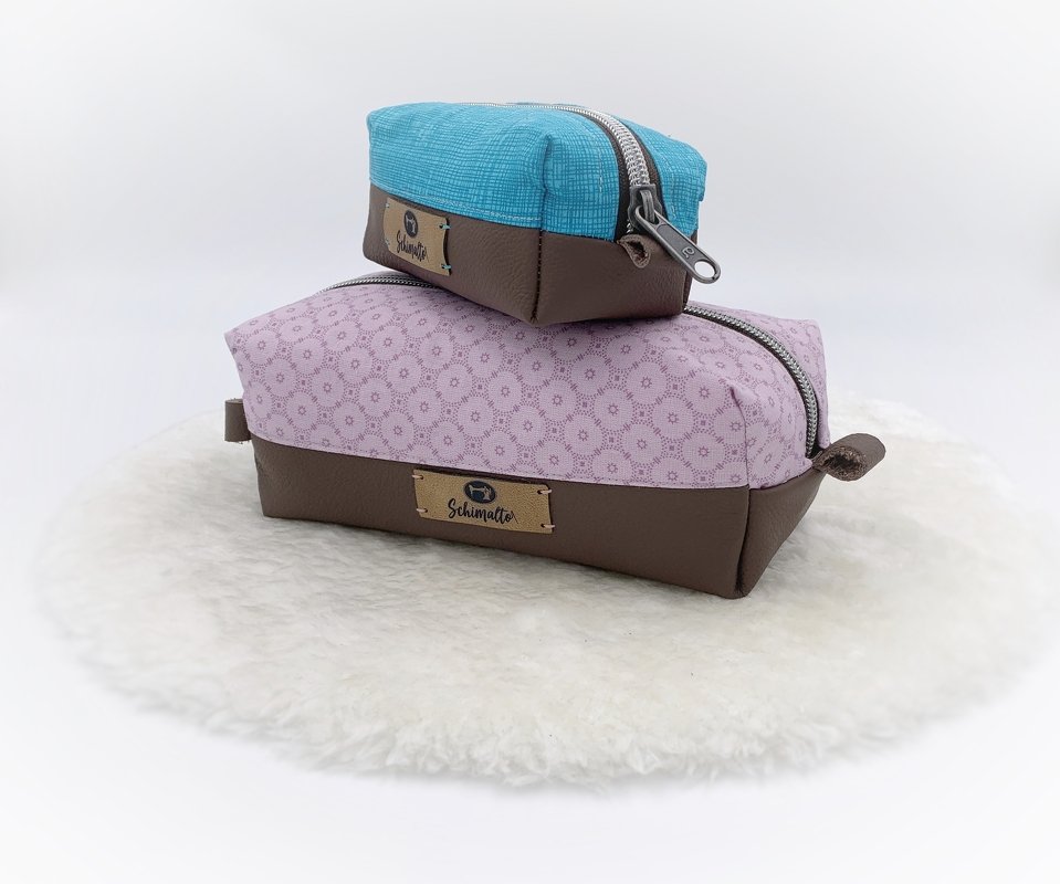 Zwei rechteckige Kosmetiktaschen, eine rosa gemustert und eine türkis, beide mit braunem Boden in Lederoptik und Reißverschluss, auf hellem Untergrund