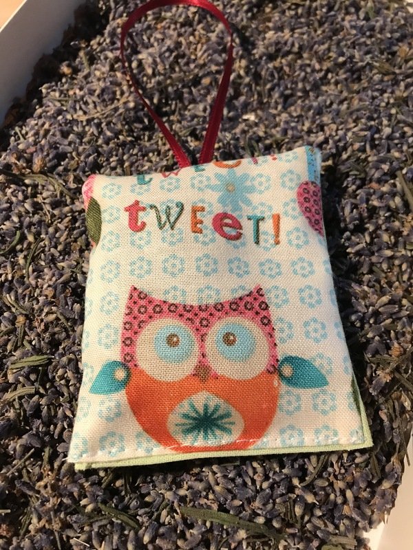Rechteckiges Lavendelsäckchen aus Baumwollstoff mit Eulenmotiv und Schriftzug „tweet!“, rotes Aufhängerband, liegt auf getrocknetem Lavendel
