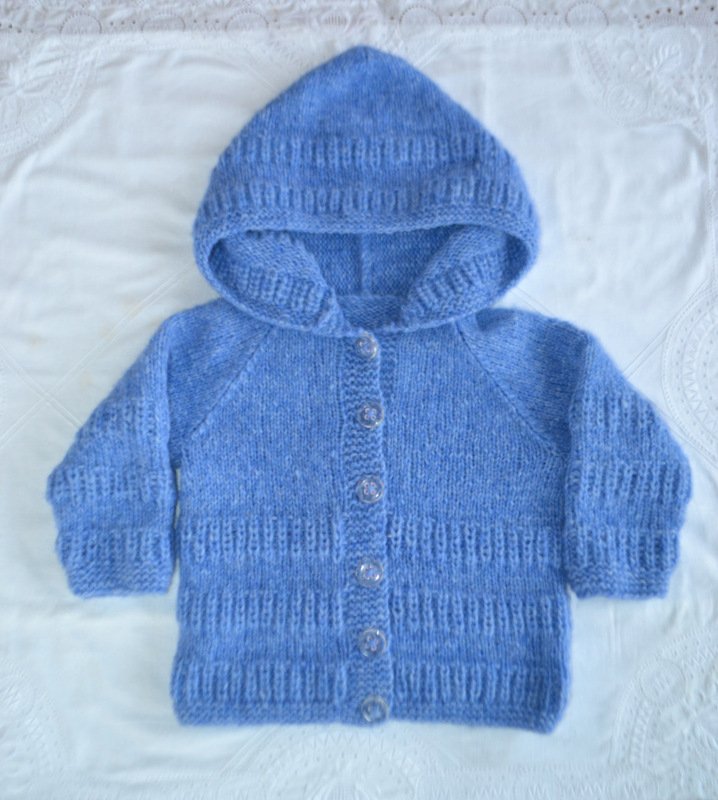 Gestrickte hellblaue Kapuzenjacke für Babys mit Knopfleiste, liegend auf hellem Stoff