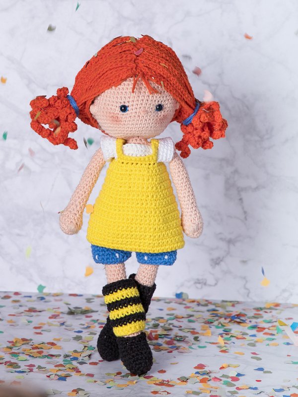 Fröhliche gehäkelte Puppe mit roten Zöpfen, gelbem Latzkleid und gestreiften Socken auf Konfetti