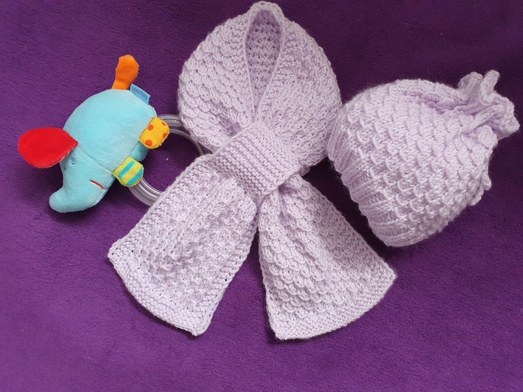 Gestrickte Babymütze und passender Schal in hellem Flieder mit Strukturmuster, daneben blaues Elefanten‑Greifspielzeug auf violettem Untergrund.