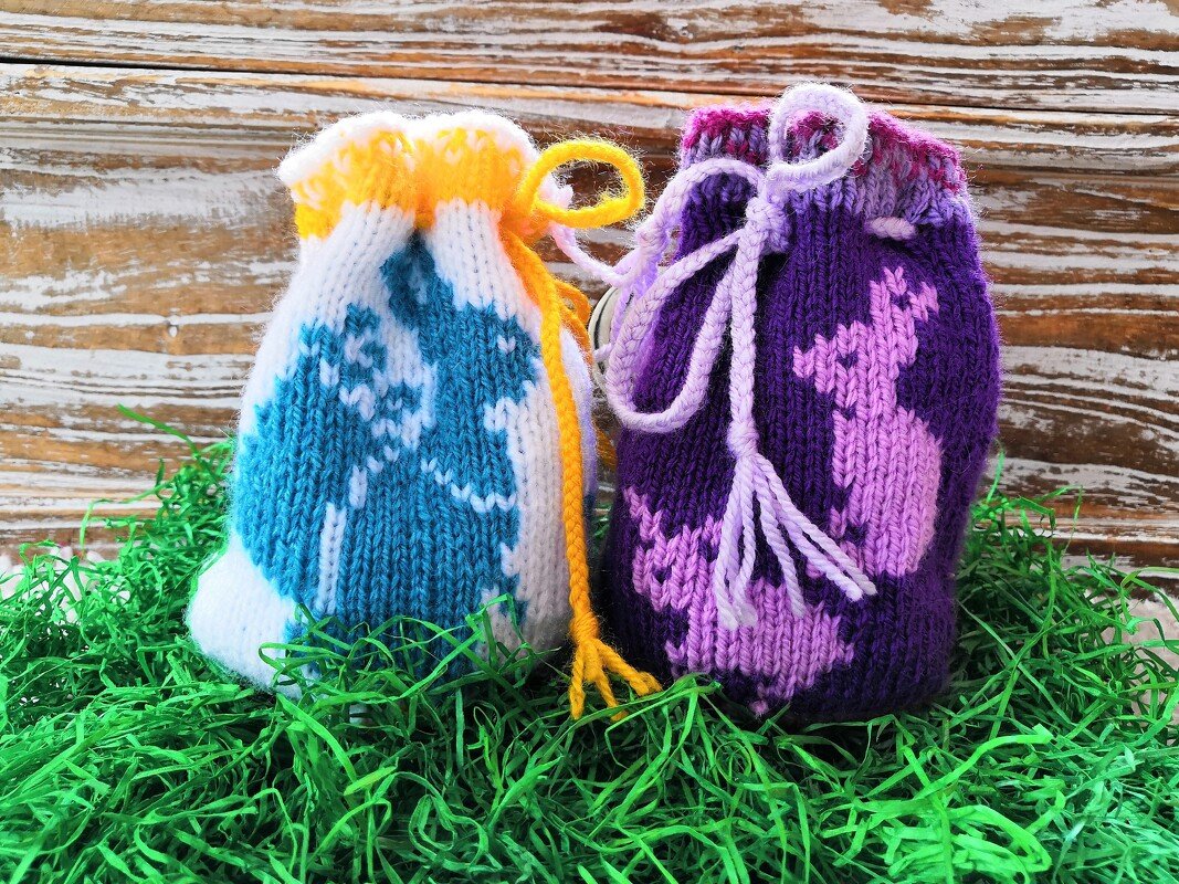Zwei gestrickte Beutel mit Hasenmuster, blau-weiß mit gelber Kordel und violett-rosa mit hellvioletter Kordel, auf grünem Ostergras vor Holzbrett