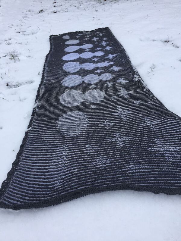 Dunkelgrauer gestrickter Schal mit hellgrauen Kreisen und Sternen auf Schnee.