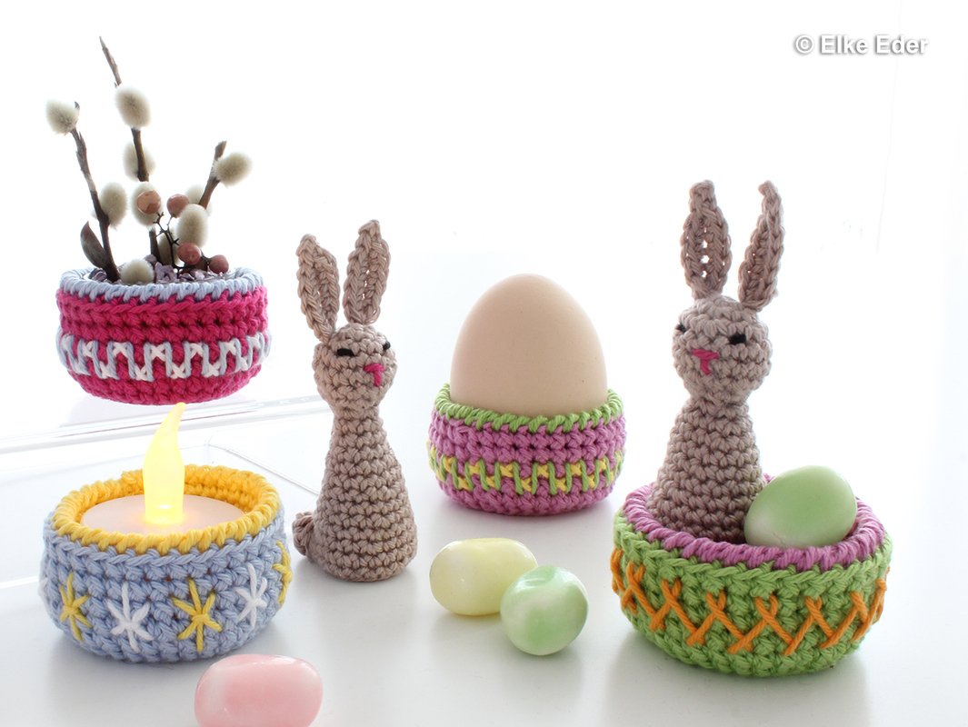 Gehäkelte Osterdekoration mit zwei Hasen, mehreren Eierwärmern, einem Teelichthalter und pastellfarbenen Deko-Eiern auf hellem Untergrund
