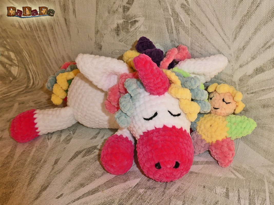 Gehäkeltes weißes Einhorn mit pinker Schnauze und pastellfarbener Mähne, liegend mit geschlossenen Augen neben kleinem Schlafengel