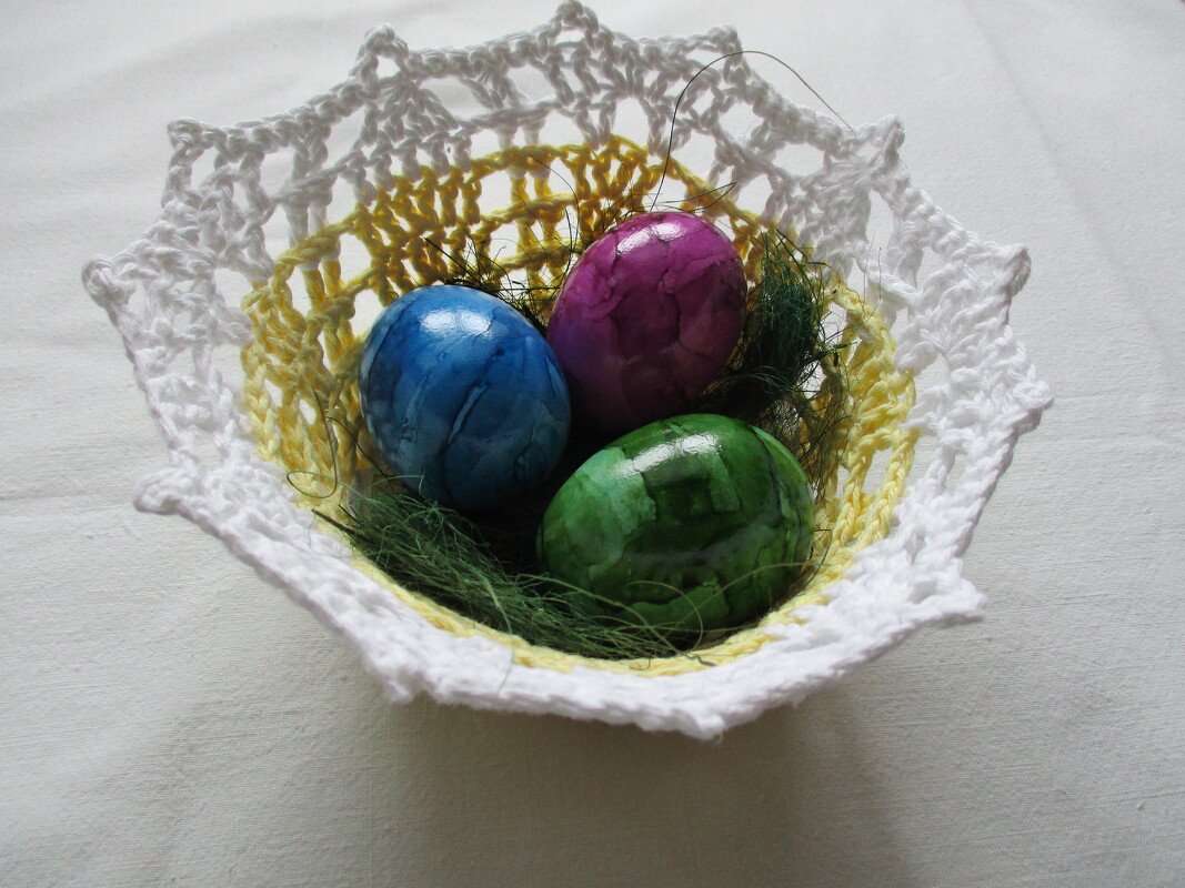 Gehäkeltes weiß-gelbes Osterkörbchen mit blauen, lila und grünen Eiern auf grünem Dekogras.