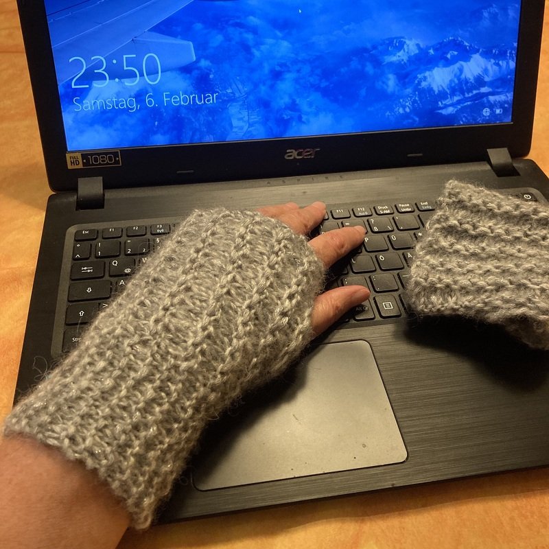 Hand mit grauem gestricktem fingerlosem Handschuh beim Tippen auf einer Laptop‑Tastatur, zweiter Handschuh liegt daneben.