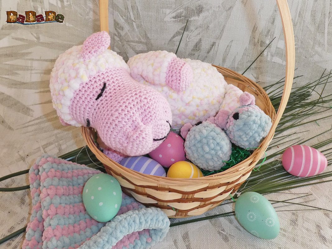 Rosa gehäkeltes Lamm und zwei kleine blaue Schäfchen in einem Weidenkorb mit bunten Ostereiern.