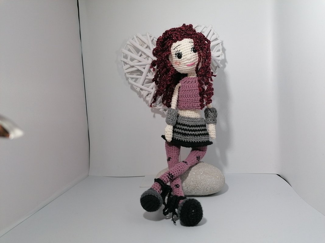 Gehäkelte Puppe sitzt auf Stein, trägt gestreiften Rock, rosa gemusterte Strumpfhosen und lockiges Haar