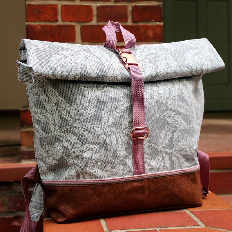 Grauer Roll-Top Rucksack mit Blättermuster, braunem Boden und rosafarbenen Gurten.