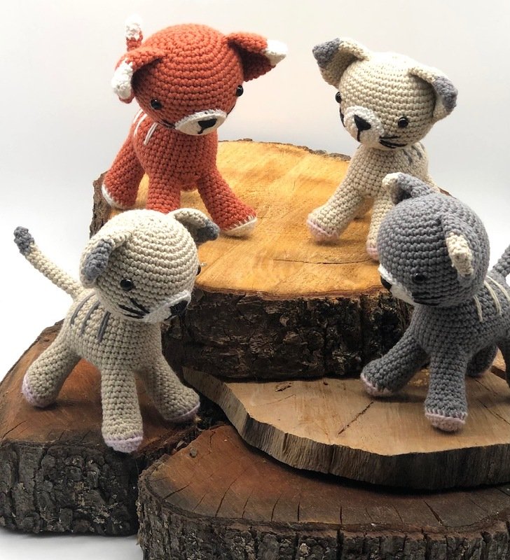 Vier gehäkelte Amigurumi-Katzen in Terrakotta, Beige und Grau auf Holzscheiben