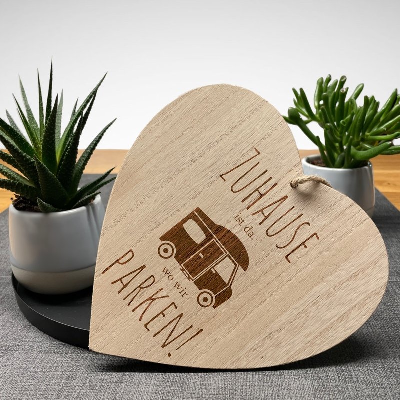 Herzförmiges Holzschild mit eingraviertem Wohnmobil und dem Spruch „Zuhause ist da, wo wir parken!“, auf Tisch mit Zimmerpflanzen