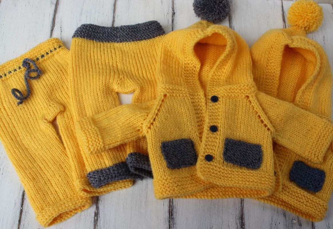 Gestricktes Babyset in Gelb und Grau mit zwei Hosen und zwei Kapuzenjacken, eine davon mit Bommel, auf hellem Holzhintergrund ausgebreitet.