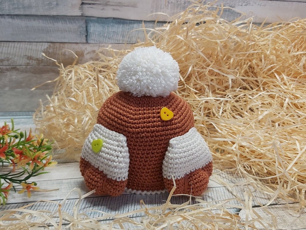 Braun-weißer gehäkelter Eierwärmer in Mützenform mit weißem Pompon und herzförmigen Knöpfen, vor Stroh dekoriert.