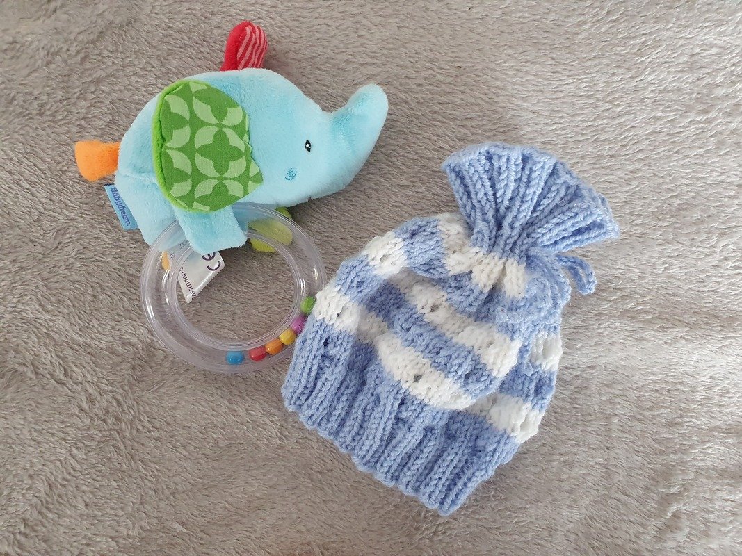 Hellblau-weiß gestreifte Babymütze aus Strick neben Rassel und Stoffelefant auf hellem Plüsch