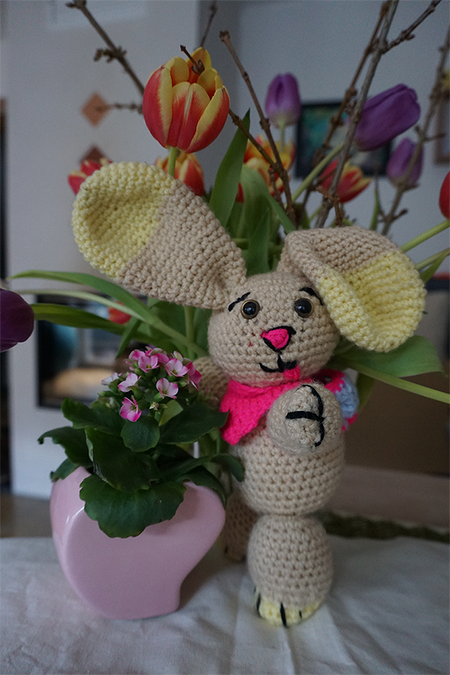 Gehäkeltes beiges Amigurumi-Häschen mit großen Ohren und pinkem Halstuch neben einem pinken Blumentopf und Tulpen