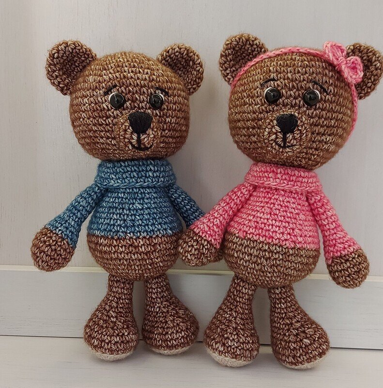 Zwei braune, gehäkelte Amigurumi-Teddybären, einer mit blauem Pullover, der andere mit rosafarbenem Pullover und Stirnband.