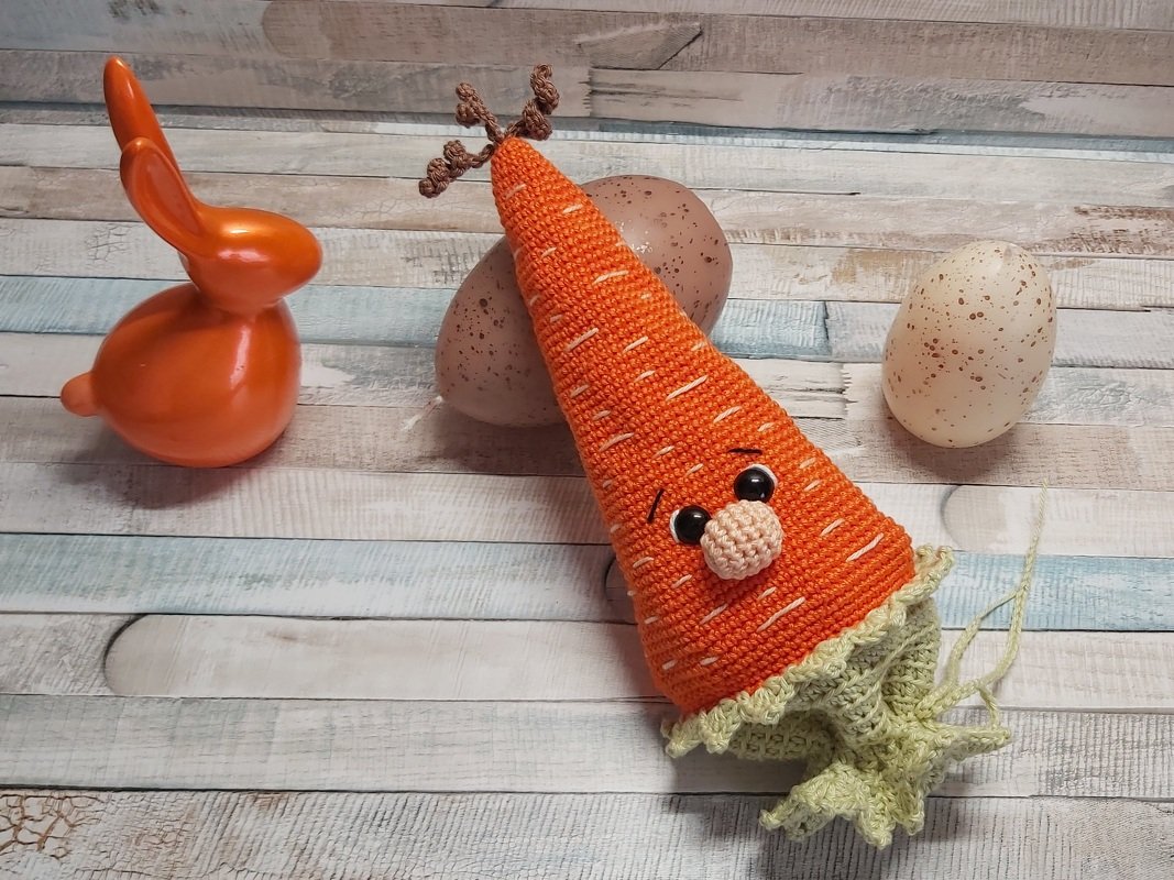 Gehäkelte orangefarbene Karotte mit Gesicht und grünen Blättern, daneben ein oranger Deko-Hase und zwei gesprenkelte Ostereier auf einem Holztisch.