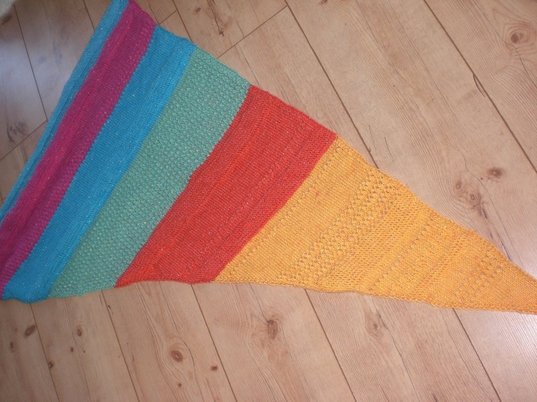 Gestricktes Dreieckstuch mit breiten Streifen (Magenta, Türkis, Mintgrün, Orange, Gelb) und feinem Glitzereffekt auf Holzboden.