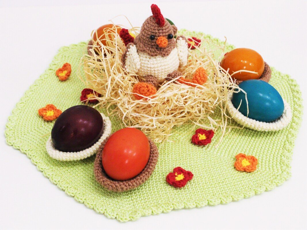 Crocheted hen in a straw nest with colorful eggs in crocheted cups on a light green scalloped mat.