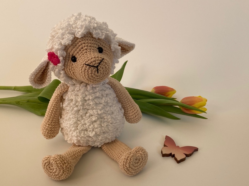 Detailansicht gehäkeltes Amigurumi-Lämmchen mit flauschigem Körper und rosa Blüte am Ohr