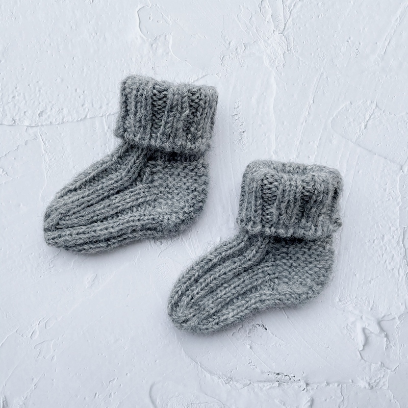 Zwei gestrickte graue Babysocken mit umgeschlagenen Bündchen auf hellem, strukturiertem Untergrund.
