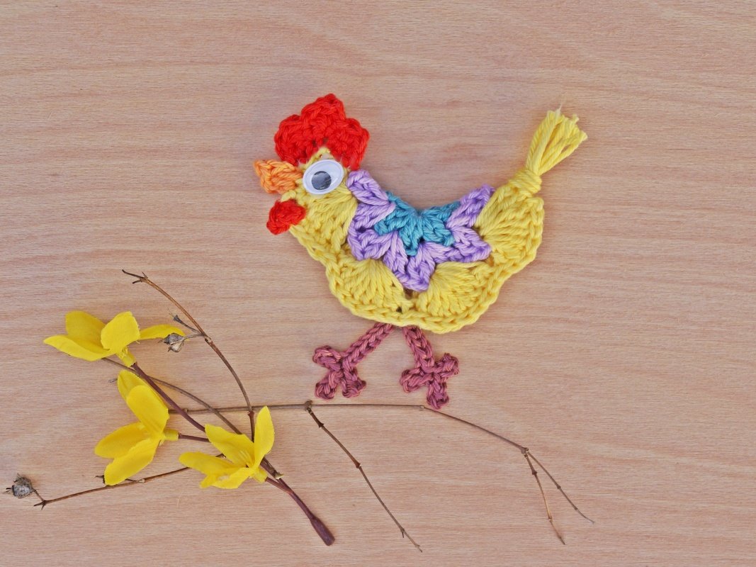 Gehäkelte gelbe Küken-Applikation mit buntem Flügelmuster und Wackelauge auf Holz neben gelben Zweigen
