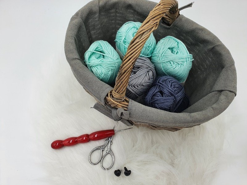 Korb mit Garnknäueln, Häkelnadel, Schere und zwei Sicherheitsaugen auf hellem Untergrund