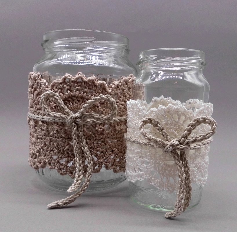 Two glass jars with crocheted sleeves—beige textured cover and white lace cover—each tied with a yarn bow on a neutral background.