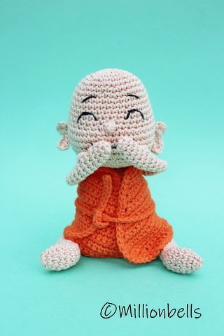 Hand-crocheted amigurumi monk-like figure in orange robe with closed eyes on teal background.