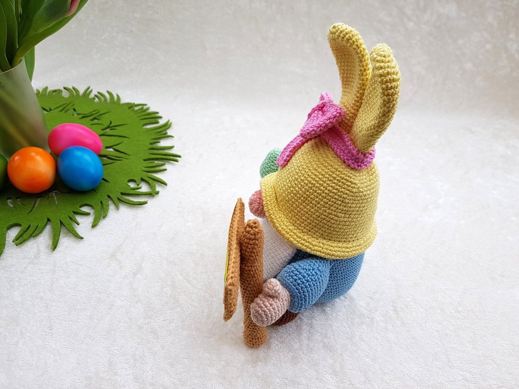 Gehäkelter Osterwichtel mit Hasenohren, gelber Mütze, rosa Schal und Karotte neben bunten Ostereiern auf grüner Unterlage