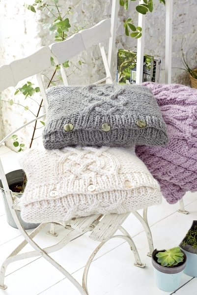 Drei gestrickte Kissenhüllen in Grau, Creme und Flieder mit Zopfmuster und Knöpfen auf weißen Stühlen