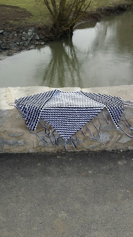 Schwarz-weißes Dreieckstuch mit Hahnentritt und Fransen auf einer grauen Steinmauer vor einem Fluss und Bäumen.