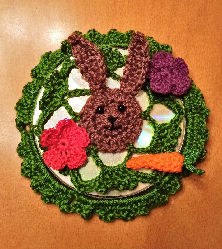 Gehäkelter Hasenkopf mit langen Ohren auf rundem grünen Häkel-Element, rosa und lila Blüten sowie orangene Karotte.
