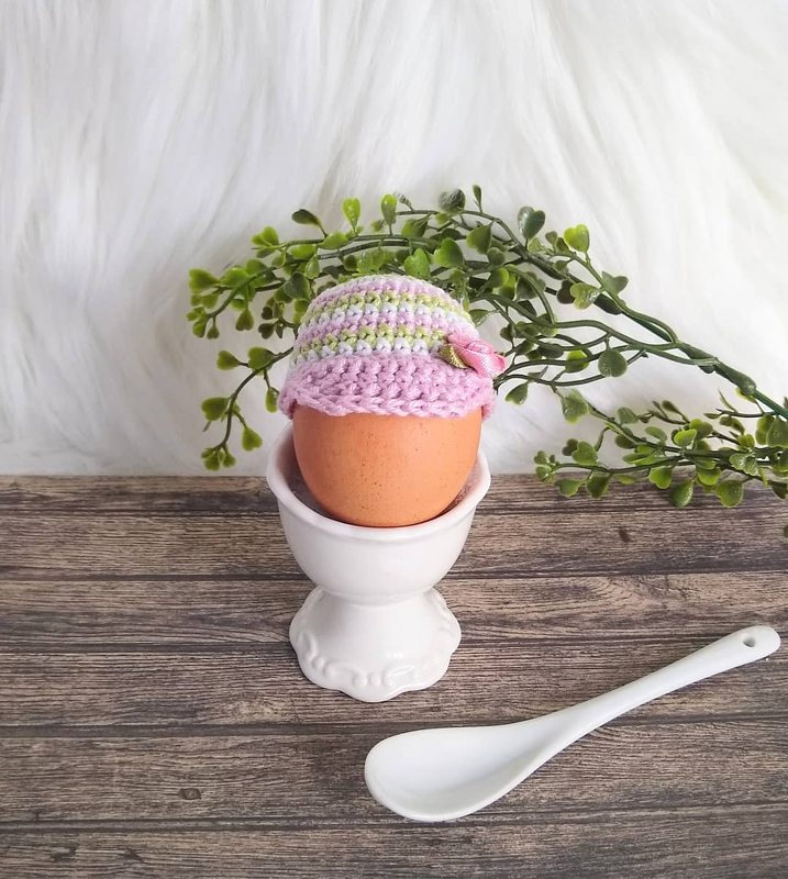 Braunes Ei im weißen Eierbecher mit gehäkeltem Eierwärmer als Schirmmütze in Pastellfarben mit kleiner Blüte